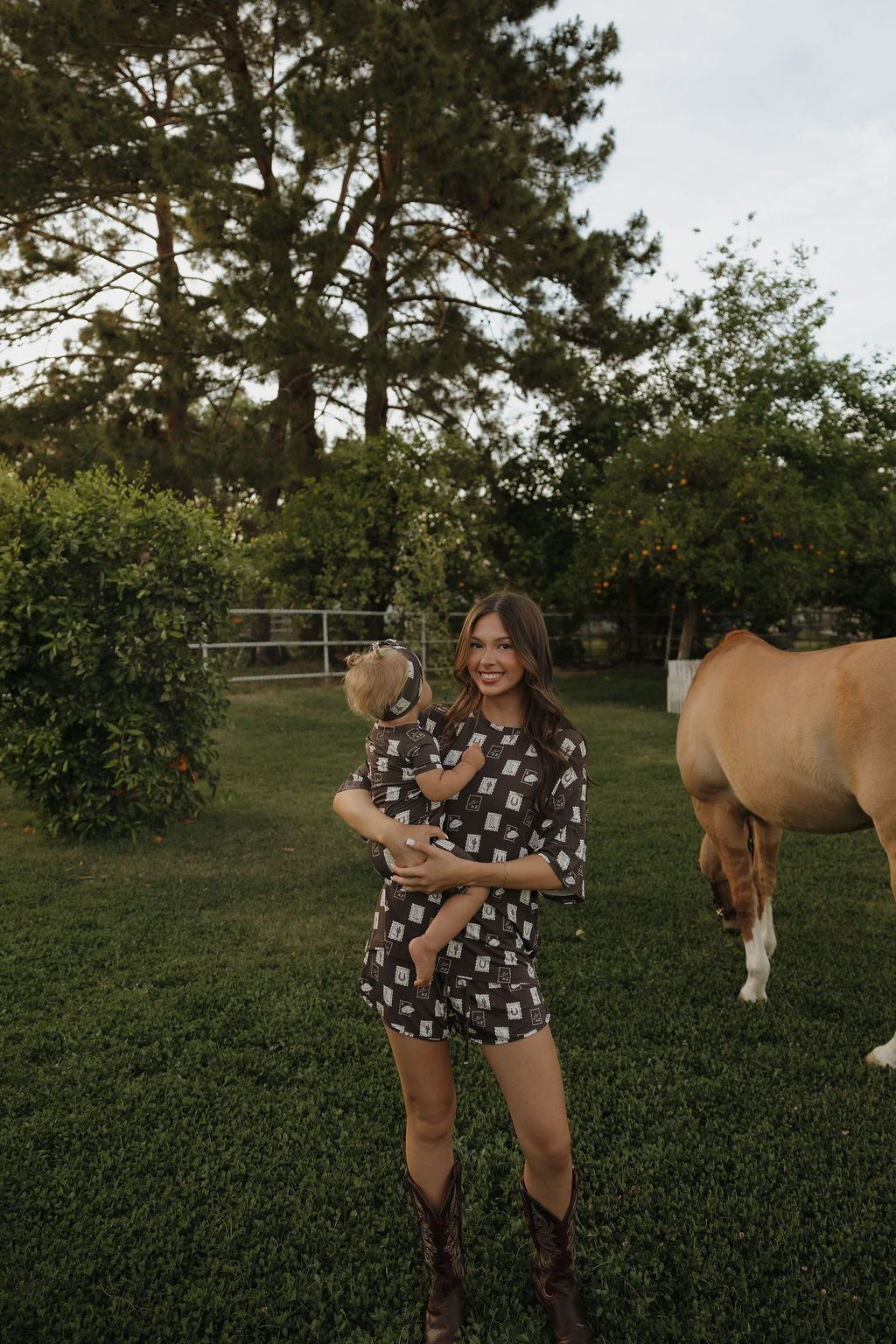 A woman in cowboy boots smiles while holding a baby on a grassy lawn, both wearing forever french baby Women's Bamboo Short Set | With Love from AZ. They are outdoors with trees, shrubs, and a brown horse grazing nearby.
