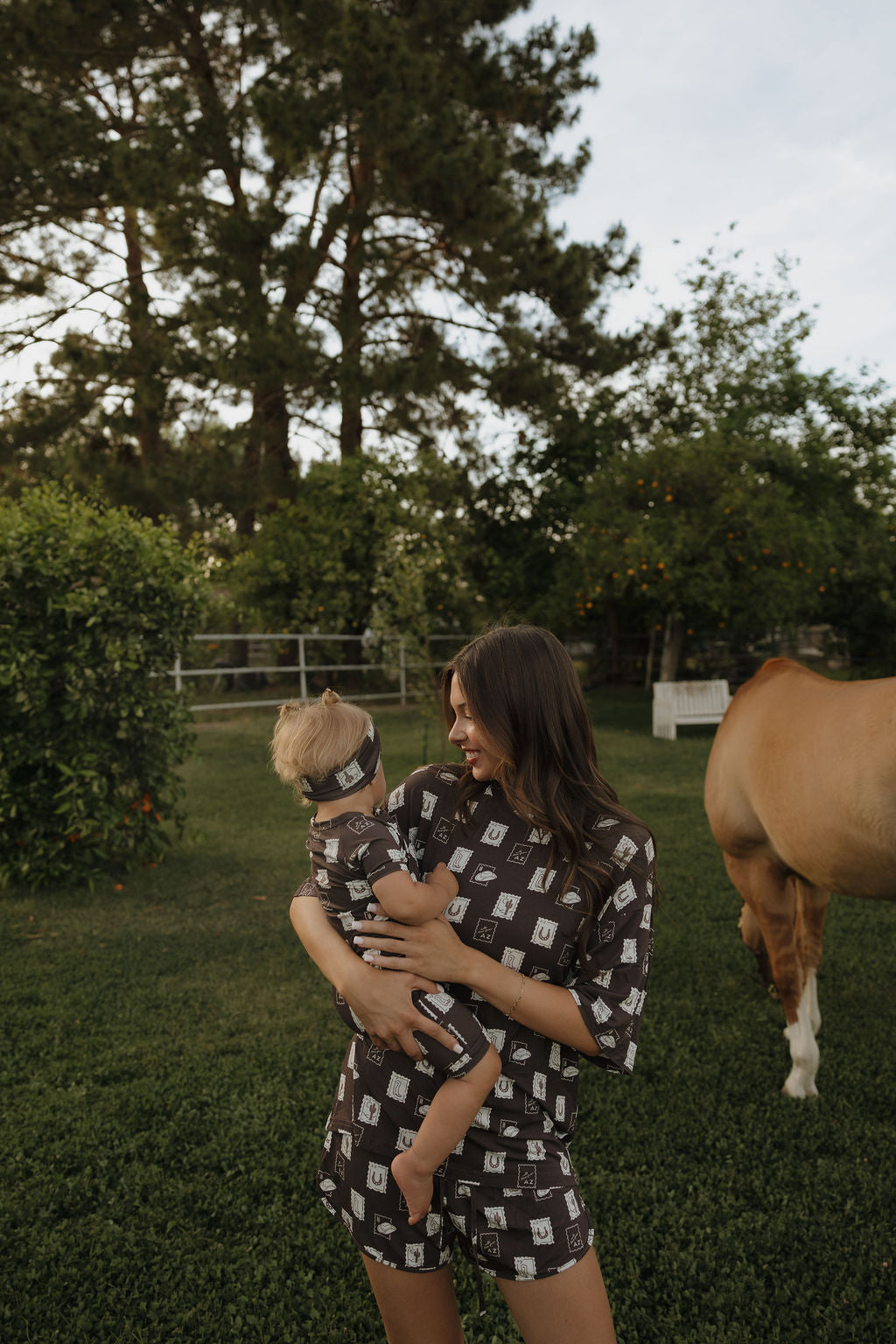 A woman in the "Women's Bamboo Short Set" by forever french baby holds a baby, both in matching breathable bamboo outfits, standing on grass near trees and a horse. The baby wears a headband.