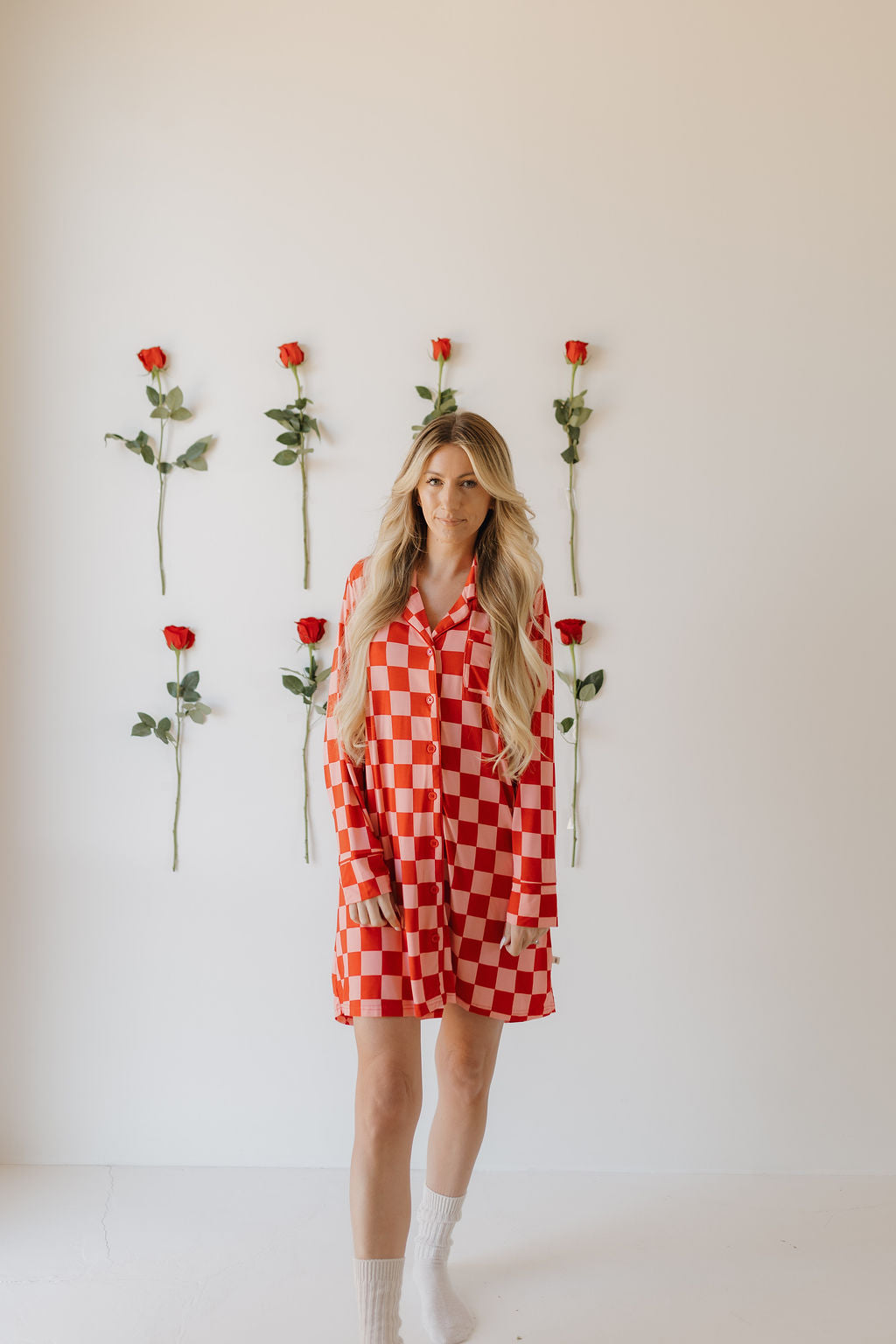 A person with long hair stands before a white wall adorned with vertical rows of red roses, wearing the eco-friendly Women's Bamboo Sleeping Dress from forever french baby in a red and white checkered pattern, paired with white socks, looking directly at the camera.