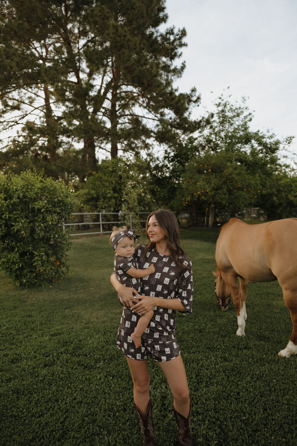 A woman in the forever french baby Women's Bamboo Short Set by With Love from AZ holds a child in matching attire, standing on grass near a horse grazing in a lush, tree-filled yard.