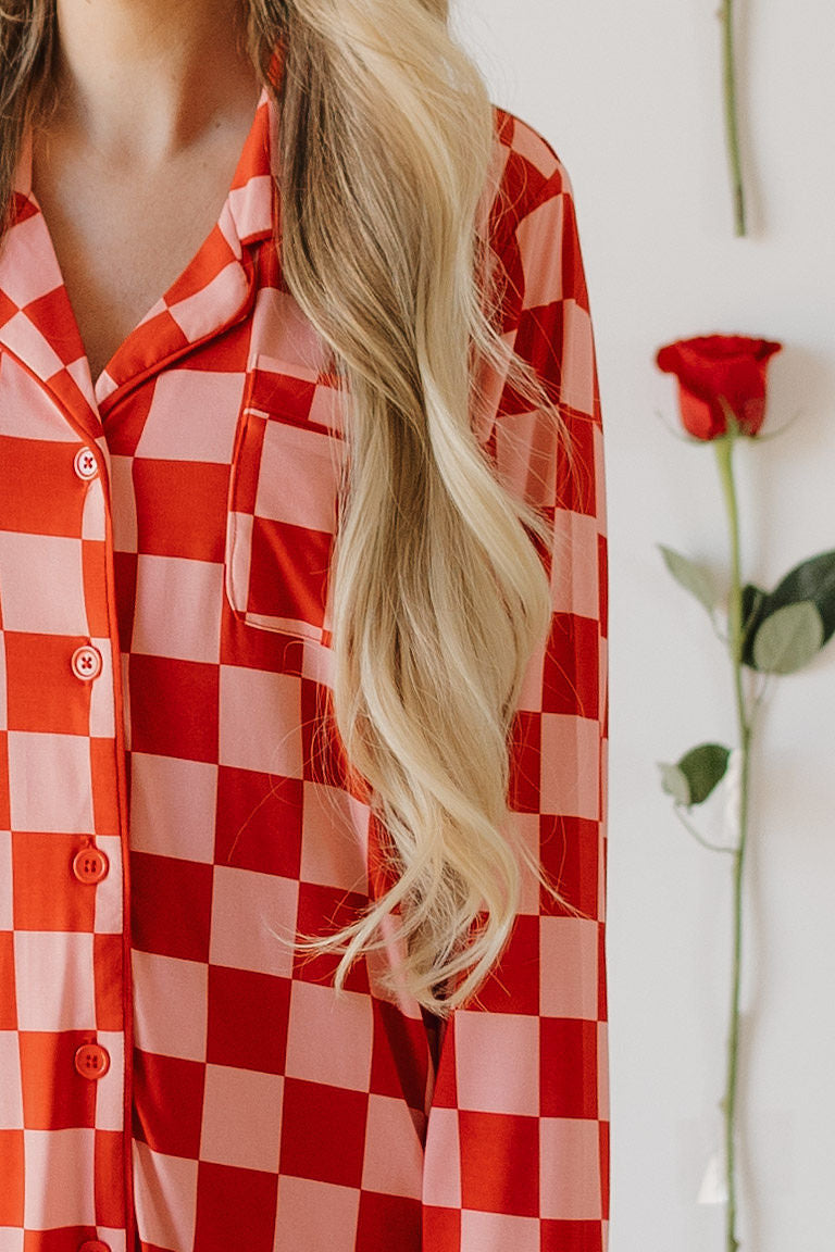 A person with long blonde hair, in a red and pink checkered Women's Bamboo Sleeping Dress by forever french baby, stands near a wall. In the background, a red rose is displayed vertically on the wall.