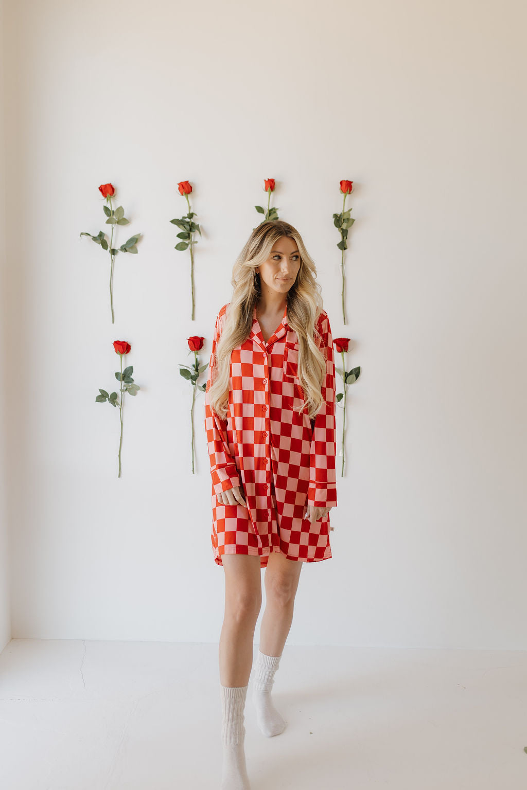 In a room with a white wall adorned by six vertical rows of red roses, someone stands wearing the Women's Bamboo Sleeping Dress | XOXO from forever french baby. The elegant red and white checkered pattern complements their long hair as they look to the side, embodying eco-friendly sophistication.