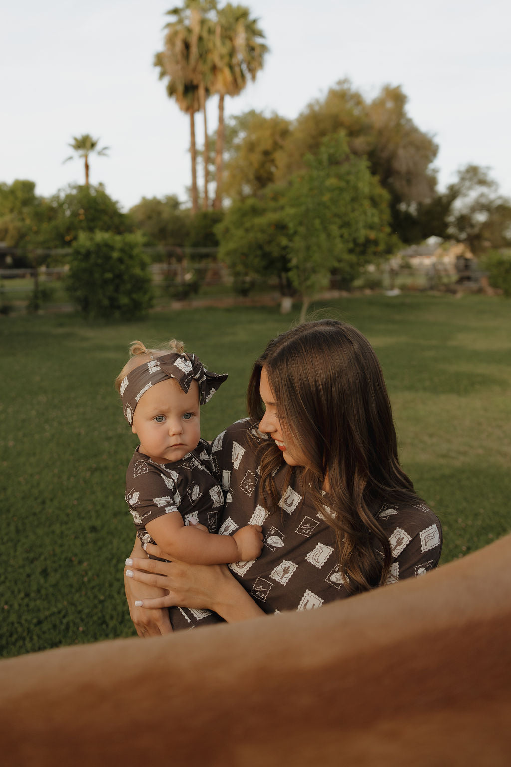 A woman holds a baby wearing the forever french baby Shortie Bamboo Zip | With Love from AZ pajamas in brown, standing on grass with trees and palm trees behind them. Both look content as a brown horse appears in the foreground.