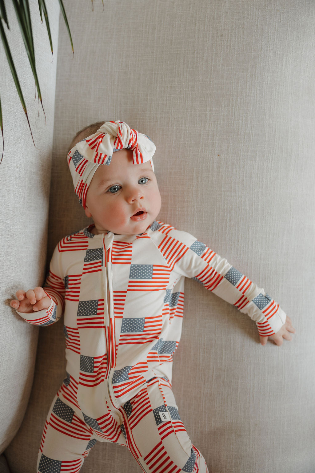 A baby in forever french baby Bamboo Zip Pajamas | Americana, featuring an American flag pattern, leans against a light cushion and looks slightly to the side.