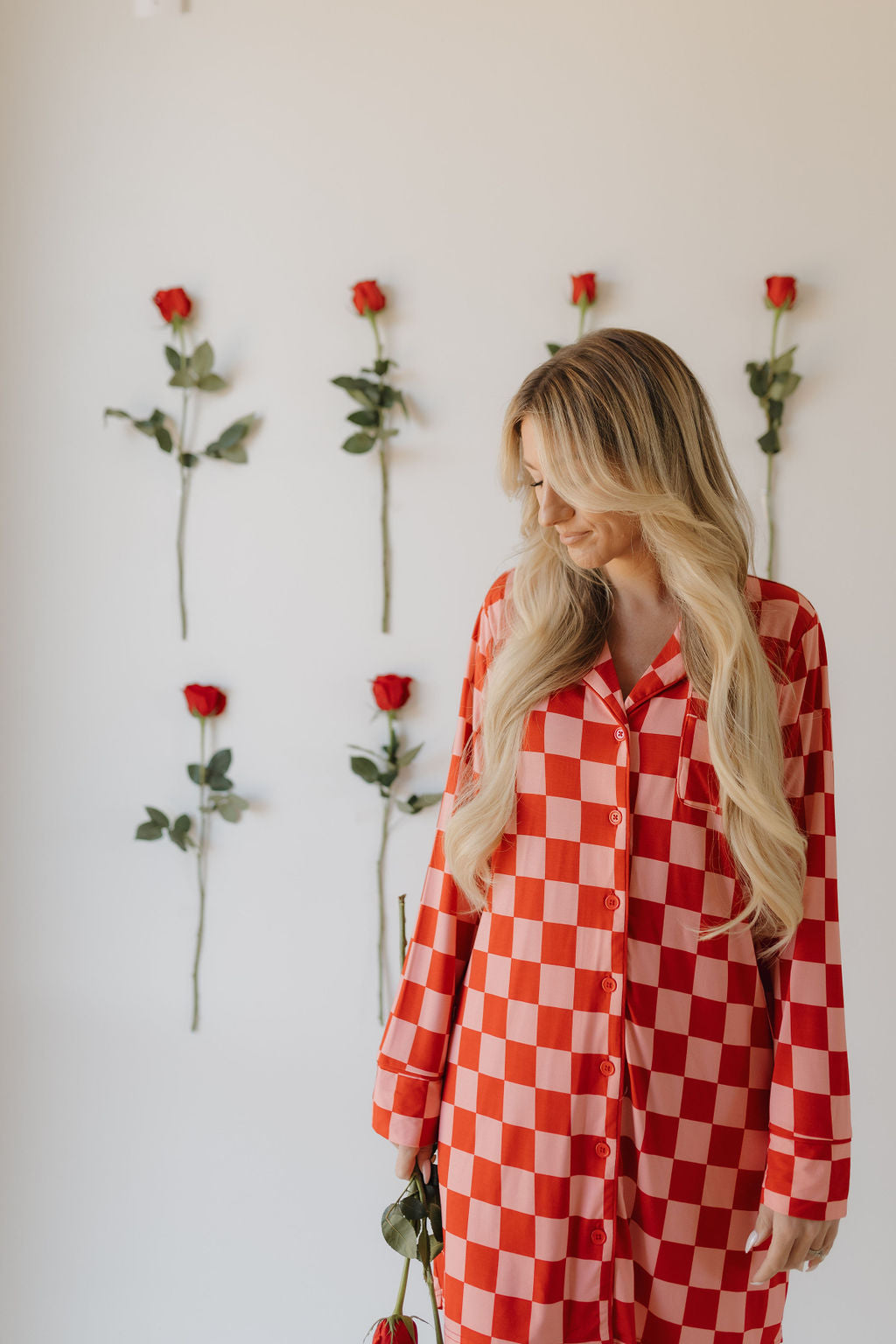 A person with long blond hair stands against a white wall decorated with vertical red roses, wearing the Women's Bamboo Sleeping Dress | XOXO by forever french baby. They hold a single rose and look downward.