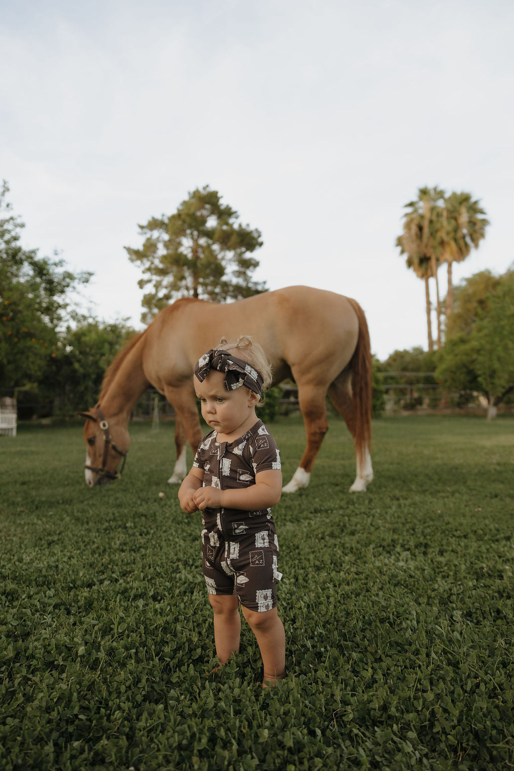 A young child in forever french baby’s Shortie Bamboo Zip | With Love from AZ pajamas and matching headband stands on green grass, looking serious, with a tan horse and trees in the background.