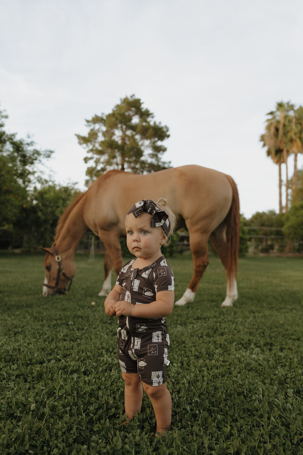 A toddler wears a forever french baby Bamboo Head Wrap | With Love from AZ, standing on grass with arms crossed and looking serious, while a tan horse grazes in the green, tree-filled yard in the background.