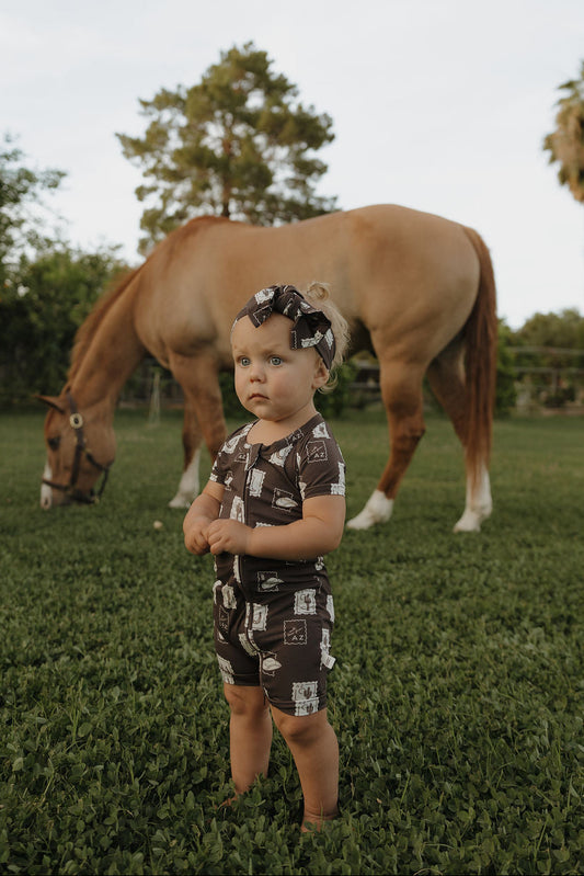 A young child wears the forever french baby Shortie Bamboo Zip | With Love from AZ—an adorable printed brown outfit with matching headband—standing seriously on grass as a tan horse grazes among trees in the background.