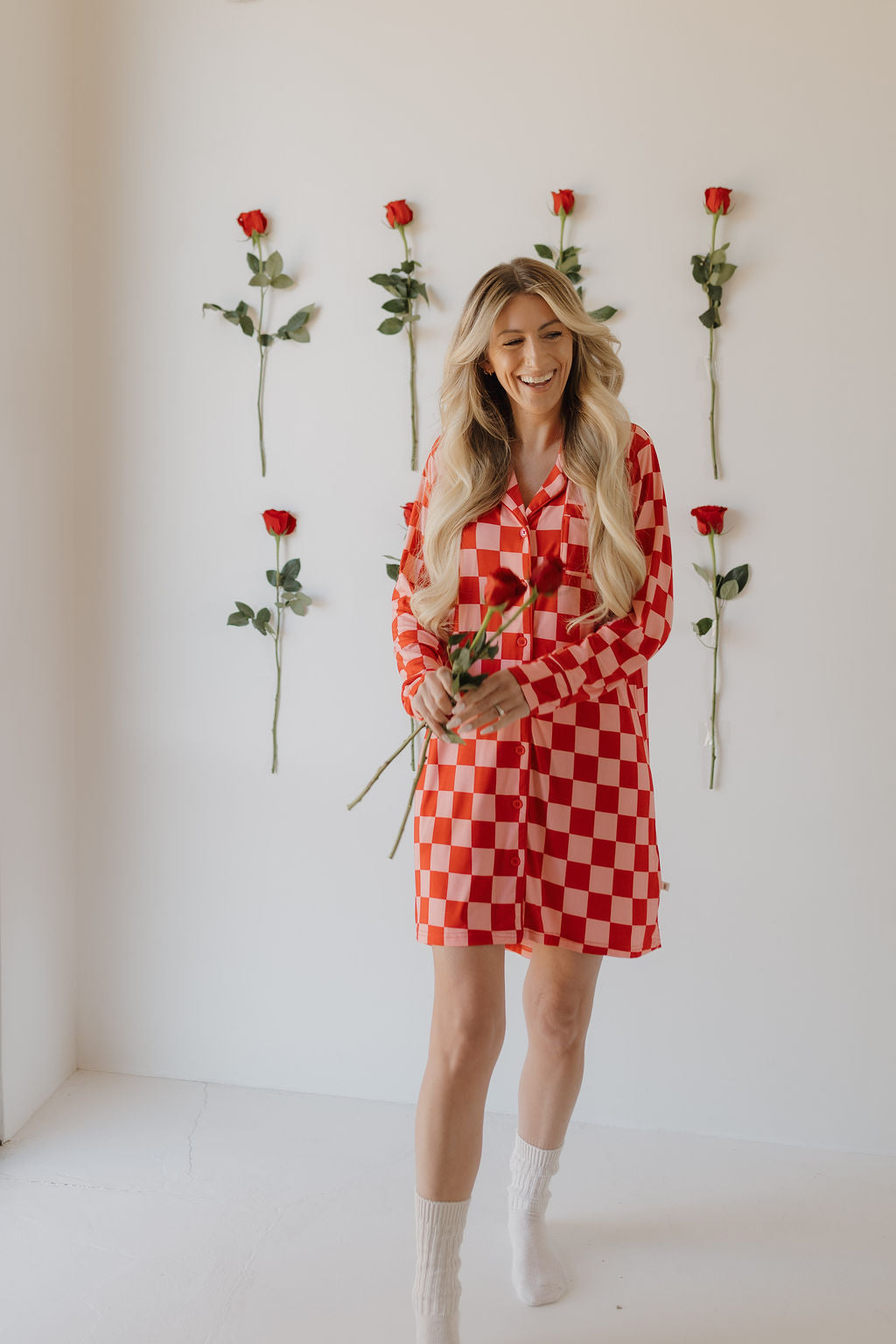 A person with long blonde hair wears a red and white Women's Bamboo Sleeping Dress by forever french baby, holding roses and smiling. They stand before a white wall adorned with vertically arranged red roses.