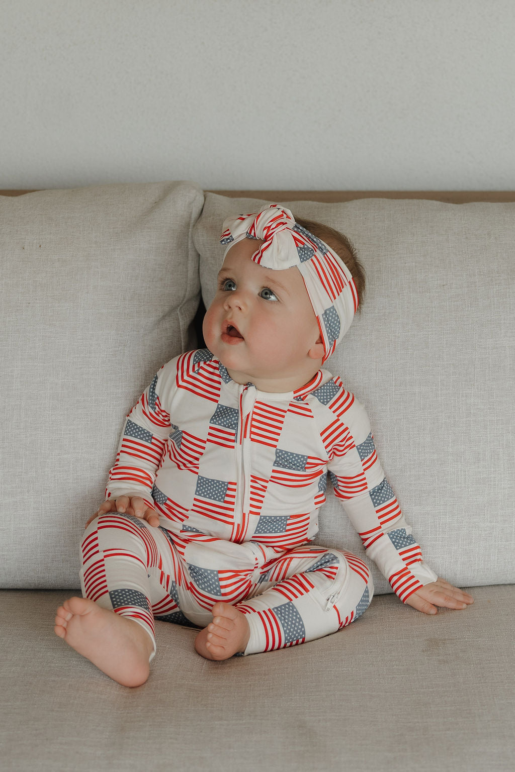 A baby in an Americana-patterned Bamboo Head Wrap by forever french baby sits on a beige couch, looking upward with curiosity.