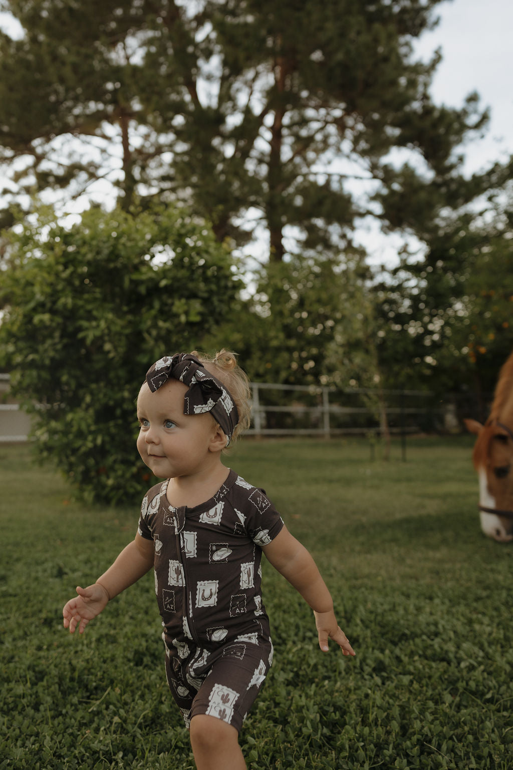 A toddler smiles while walking on grass in a yard, wearing the forever french baby Shortie Bamboo Zip | With Love from AZ pajamas, with a matching headband, as trees and a horse grazing appear in the background.