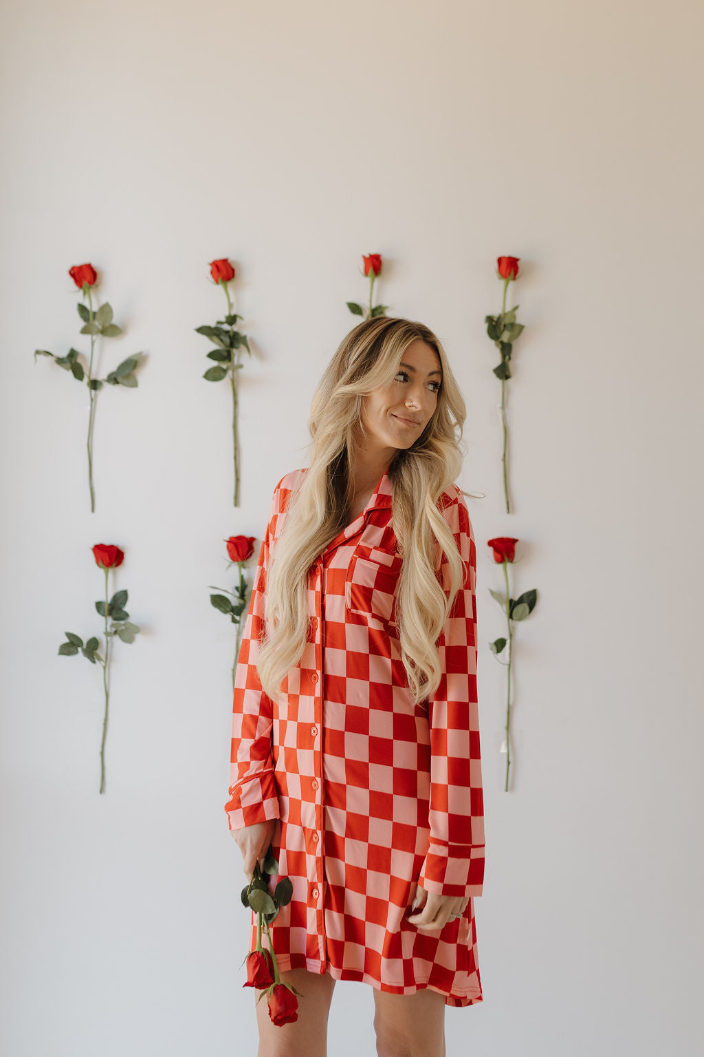 A woman with long blonde hair, dressed in a Women's Bamboo Sleeping Dress by forever french baby, stands holding a rose. Behind her, eco-friendly décor features red roses aligned vertically on the white wall, adding sustainable beauty.