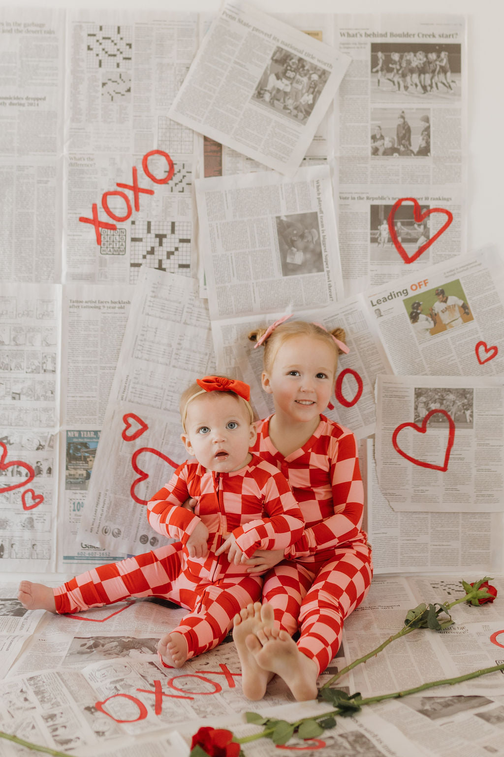 Two children in "forever french baby" Bamboo Zip Pajamas | XOXO in red and white checkered pattern sit on a newspaper-covered floor. The older child smiles, hugging the younger one with red hearts, "XOXO" art, and roses scattered around, forming a cozy scene.