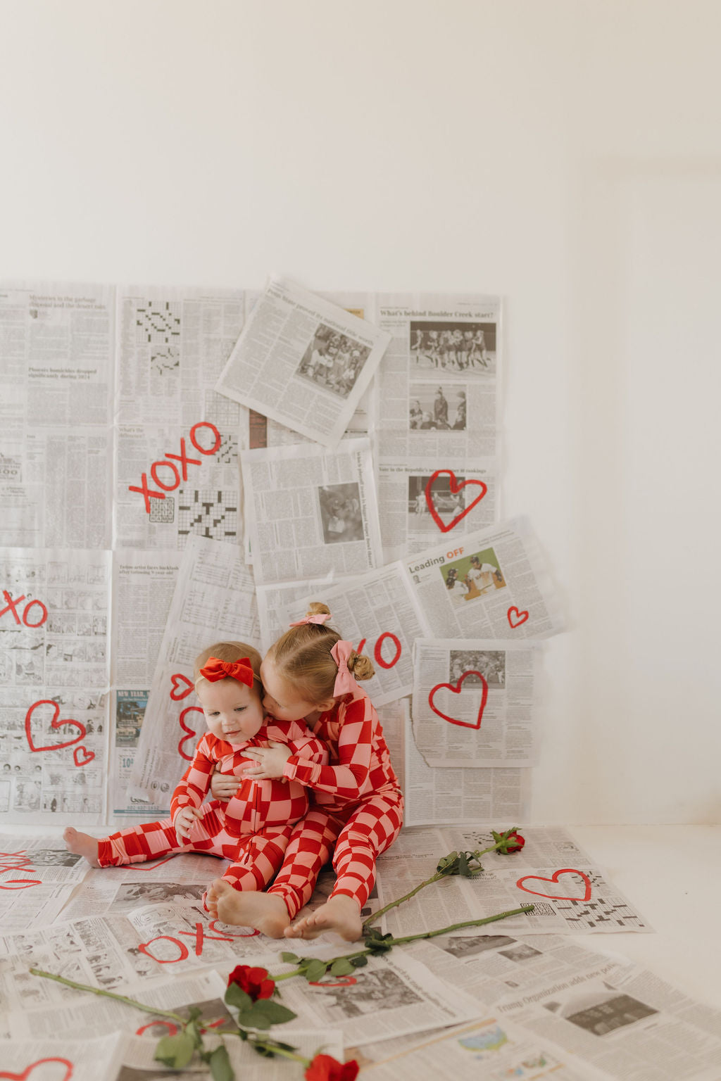 Two children in matching forever french baby Bamboo Two Piece Pajamas | XOXO sit on a floor covered with newspapers. The older child hugs the younger one, cozy in breathable fabric. Red hearts and "XOXO" are drawn on the newspaper wall, with roses scattered around them.