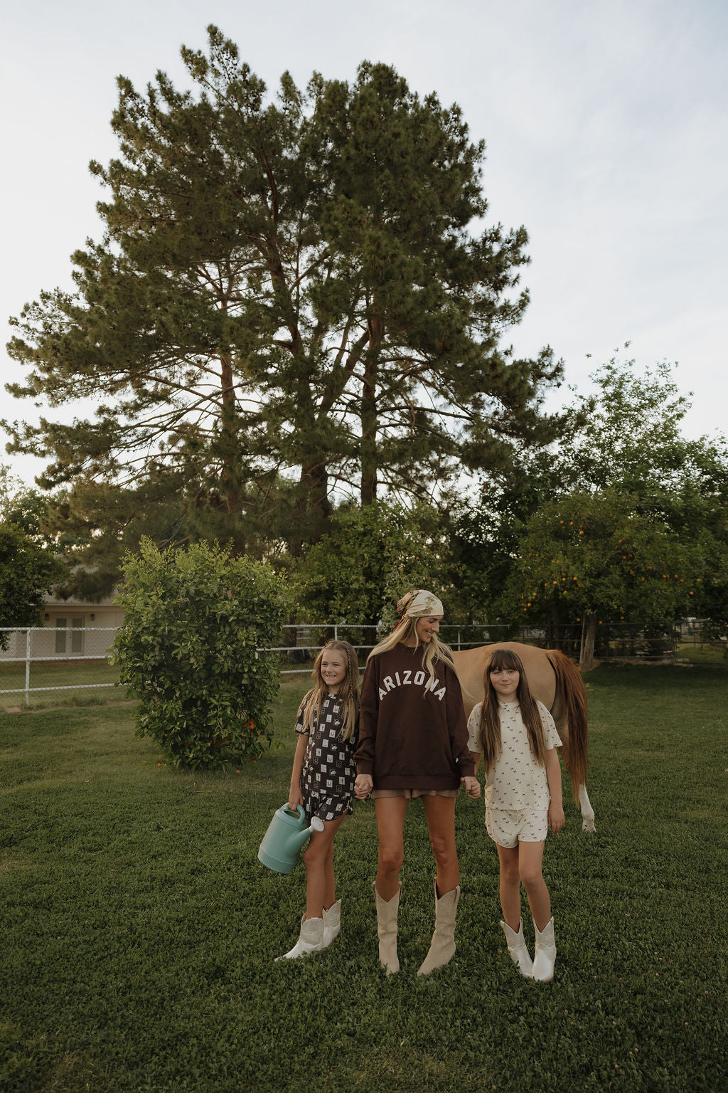 A woman and two girls in cowgirl boots and forever french baby's Girl's Bamboo Short Set | With Love from AZ stand on grass by a horse and pine tree. One girl holds a watering can in their fenced yard with trees and a house behind.