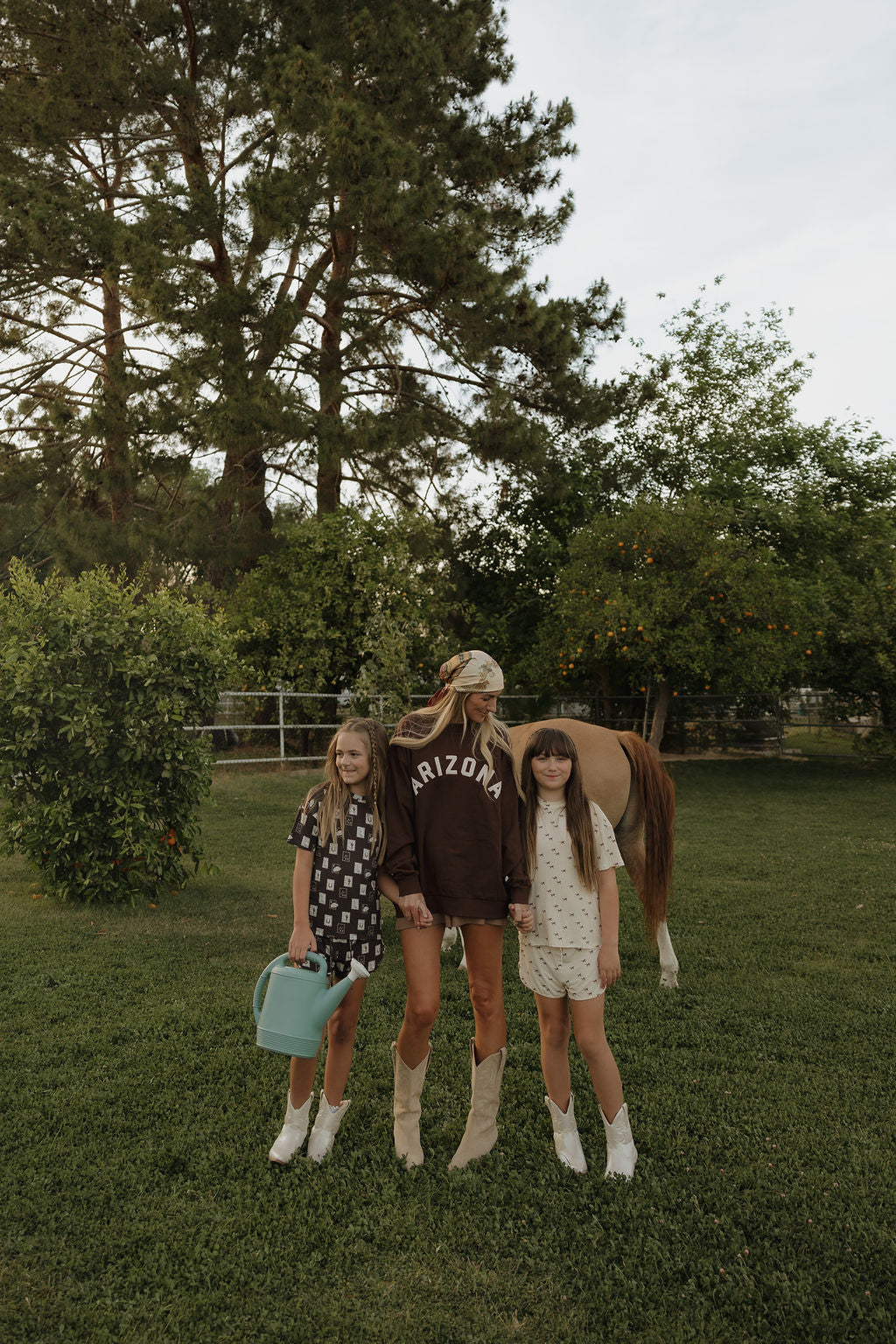 A woman and two girls, dressed in forever french baby’s Girl's Bamboo Short Set | Wild Horses, stand on grass by a horse. One girl holds a blue watering can; the woman’s sweatshirt says “ARIZONA,” with trees and a fence in the background.
