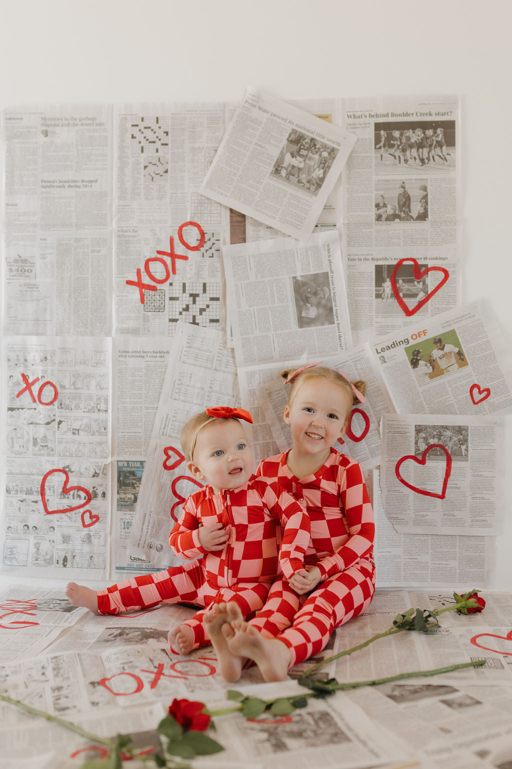 Two young children in matching Bamboo Two Piece Pajamas | XOXO by forever french baby sit on a newspaper-covered floor. The backdrop includes red hand-drawn hearts and "XOXO" symbols, with a single red rose on the floor. Both kids smile, enjoying their soft, breathable attire.