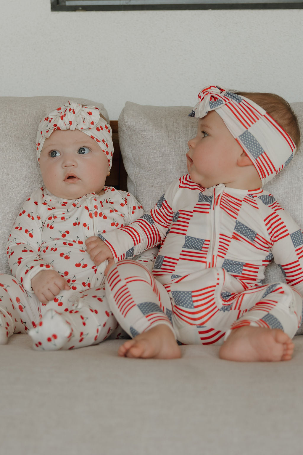 Two babies sit on a light couch. One wears a cherry outfit with matching head wrap, while the other looks over in a stars-and-stripes outfit and the forever french baby Bamboo Head Wrap | Americana.