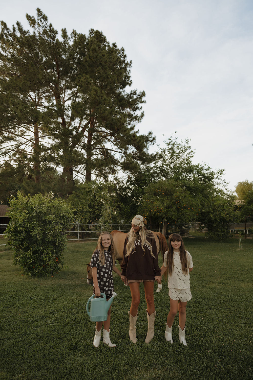 Two girls and a woman in western outfits and boots stand on green grass. One girl, wearing the Girl's Bamboo Short Set by forever french baby, holds a watering can. A horse stands behind them with trees in the background.