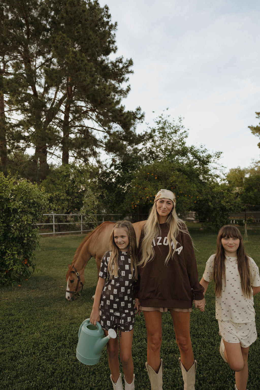 A woman in a relaxed-fit Adult Sweatshirt | ff Arizona by forever french baby stands on grass holding hands with two young girls, one holding a blue watering can. A horse grazes nearby amid lush trees and a fence in this serene outdoor scene.