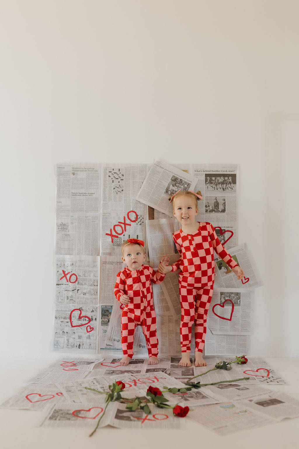 Two children wearing forever french baby's breathable Bamboo Two Piece Pajamas in a red and white checkered pattern stand on whimsical newspapers displayed with red hearts and "XOXO" doodles. They hold hands amidst scattered roses.