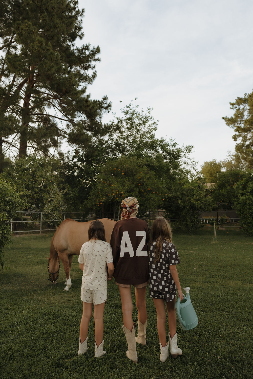 Three people stand on grass facing away: an adult in a relaxed fit Adult Sweatshirt | ff Arizona by forever french baby, and two children, one holding a blue watering can. A brown horse grazes nearby among tall trees. The sweatshirt is 100% cotton.