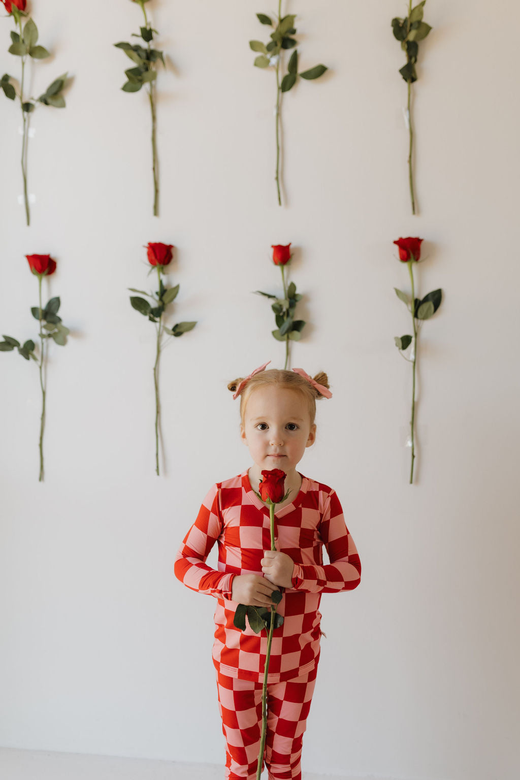 A child in forever french baby's Bamboo Two Piece Pajamas | XOXO, in a red and white checkered design made from breathable fabric, holds a red rose against a white wall decorated with vertical rows of roses. Their two small buns frame their face as they gaze at the camera.