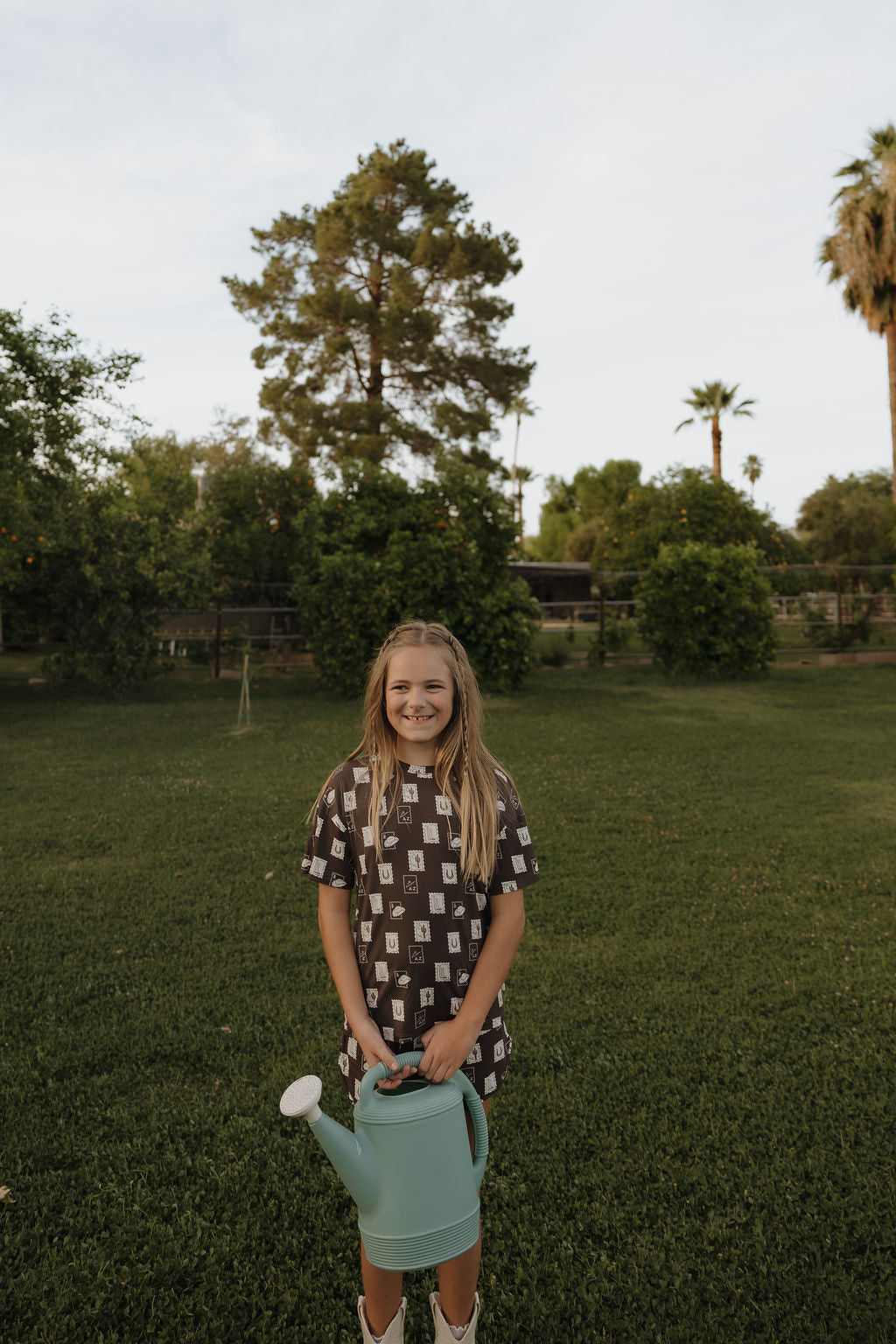 A young girl in the Girl's Bamboo Short Set by forever french baby smiles on a grassy lawn, holding a blue watering can amid trees and bushes under a partly cloudy sky.