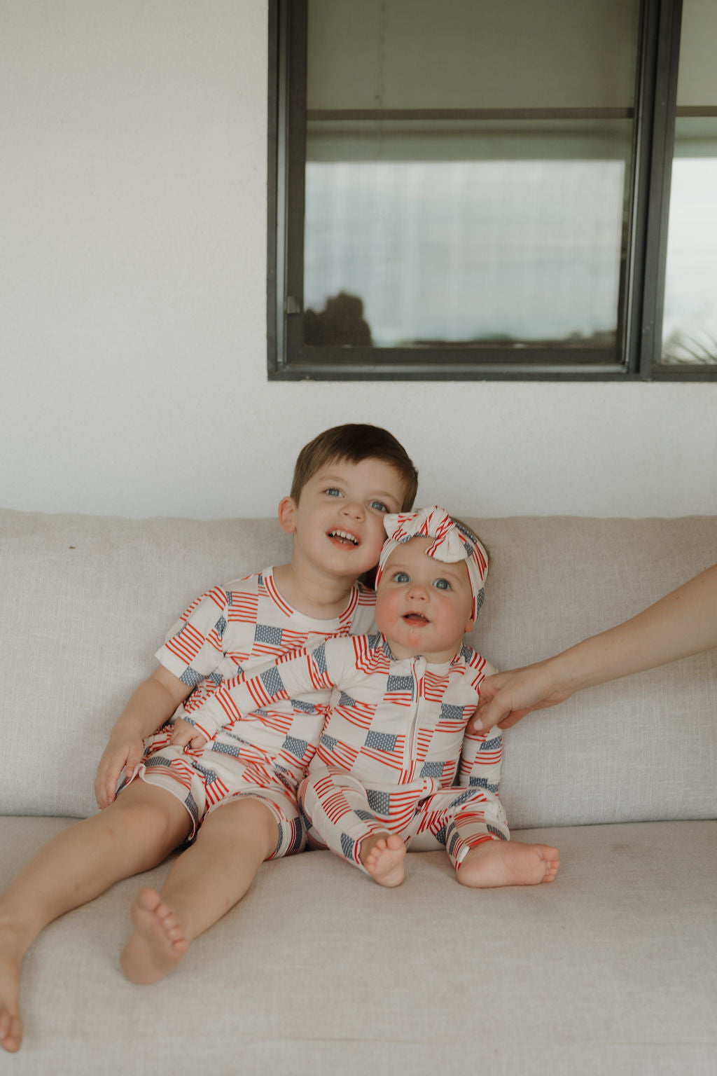 Two young children, a boy and a baby girl, sit on a light couch wearing forever french baby Bamboo Zip Pajamas | Americana featuring red, white, and blue flag prints. The hypo-allergenic pajamas keep them cozy as an adult supports the baby. A window is behind them.