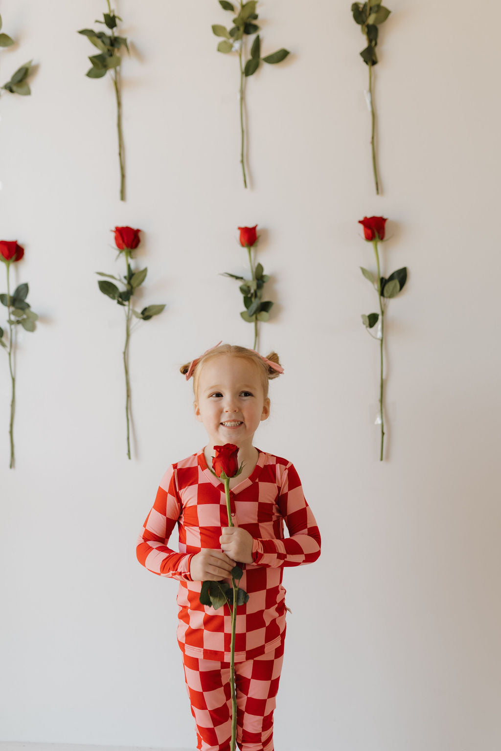 A young child, wearing forever french baby's Bamboo Two Piece Pajamas in red and white checkered pattern, holds a red rose. The background shows several vertically arranged red roses on a white wall. The child smiles at the camera.