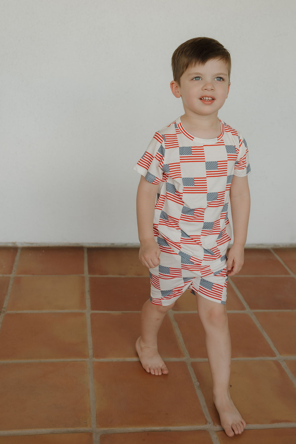 A young boy with short brown hair wears the forever french baby Bamboo Short Set | Americana, featuring a red, white, and blue flag pattern, as he stands barefoot on terracotta tiles against a plain white wall.