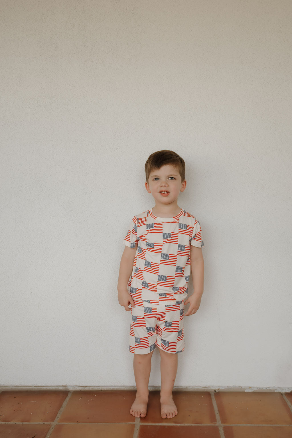 A young boy stands barefoot on terracotta tiles against a plain white wall, wearing the forever french baby Bamboo Short Set | Americana—breathable bamboo t-shirt and shorts featuring a red, white, and blue geometric flag pattern.