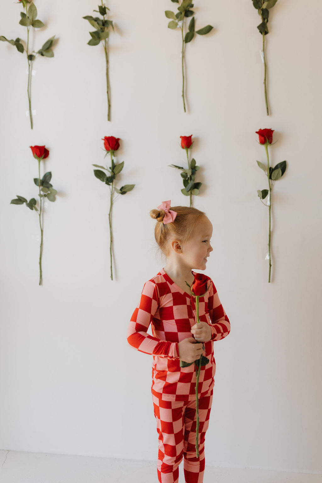 A young child stands indoors, wearing bamboo two-piece pajamas from forever french baby with a red and white checkered design. They hold a red rose and face sideways, while multiple red roses hang upside down on the wall behind them.