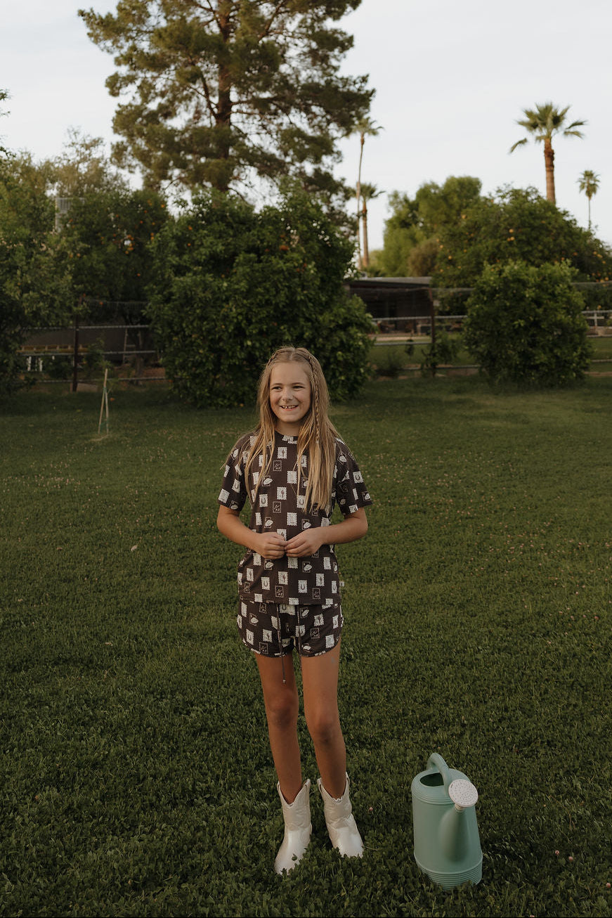 A young girl with long blond hair stands on grass in a yard, smiling in the Girl's Bamboo Short Set by forever french baby. White boots, a green watering can, trees, and a fence complete the cheerful scene.