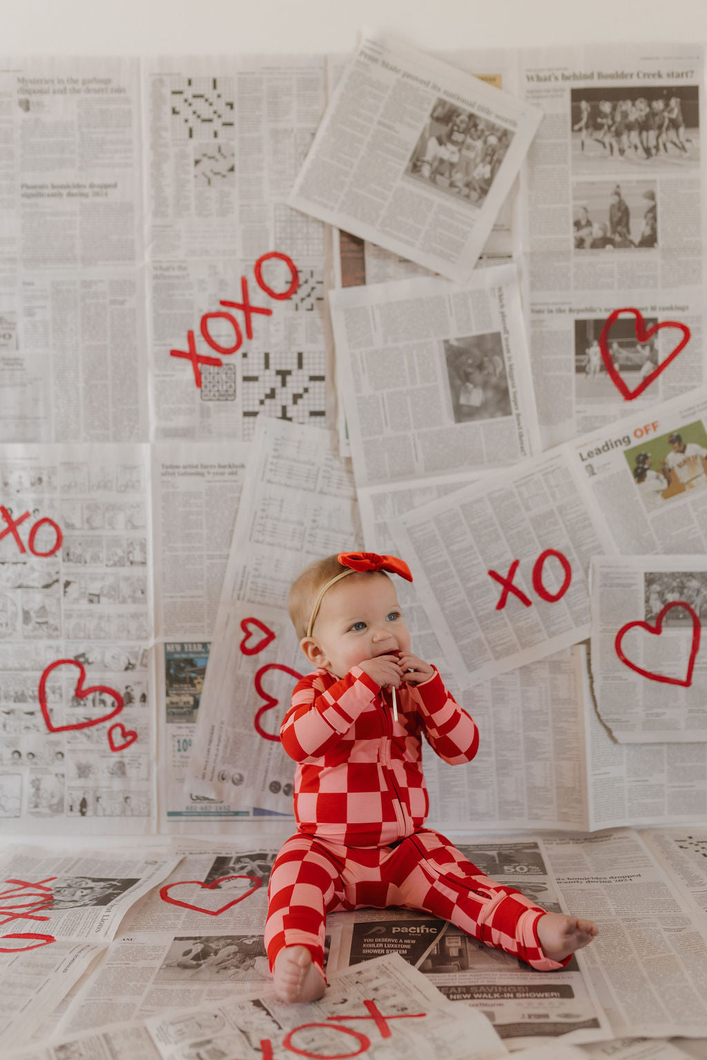 A baby in forever french baby's Bamboo Zip Pajamas | XOXO, known for being hypo-allergenic, sits on a newspaper-covered floor. The backdrop features more newspaper pages with red hearts and "XO" symbols. A matching red bow adorns the baby's head.