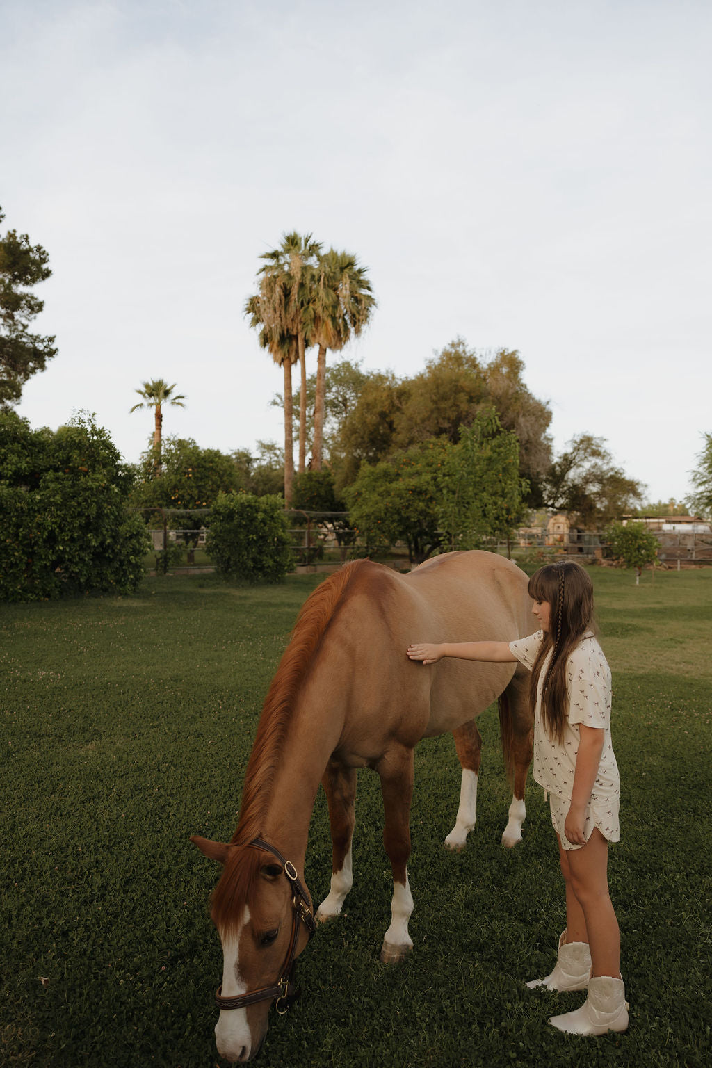 A young girl wears the forever french baby Girl’s Bamboo Short Set | Wild Horses, hypo-allergenic and perfect for sensitive skin, as she stands beside a brown horse grazing on green grass under clear skies with trees and palms in the background.