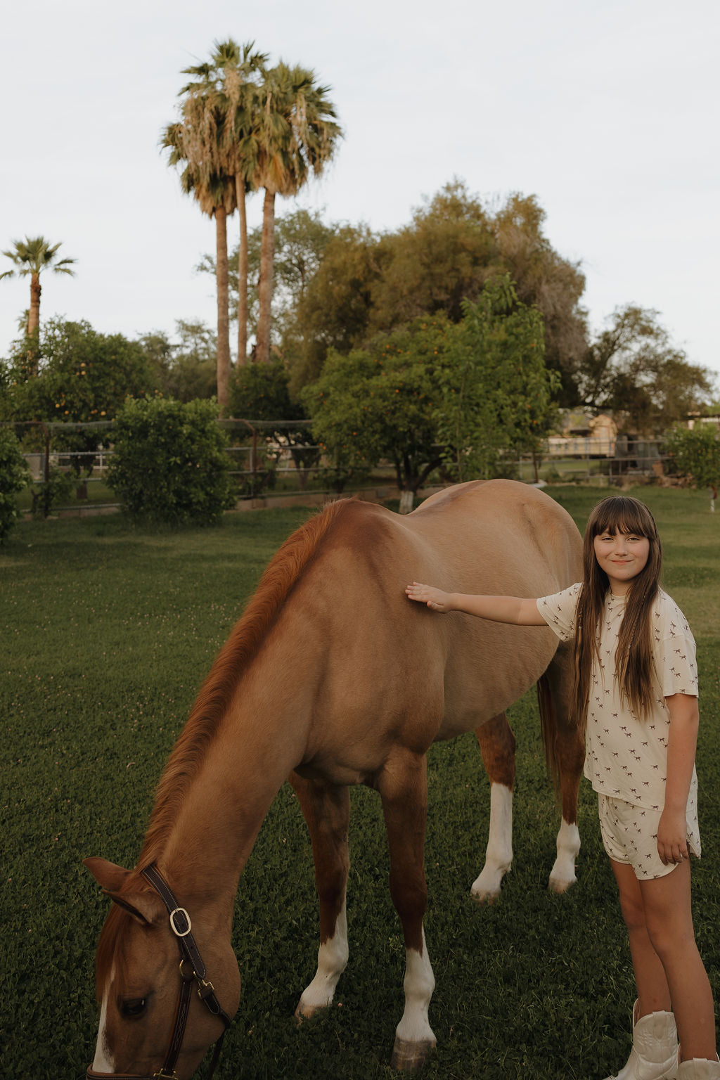 A young girl with long hair, wearing the Girl's Bamboo Short Set | Wild Horses by forever french baby and boots, stands beside a brown horse grazing in a green yard with palm trees and bushes.