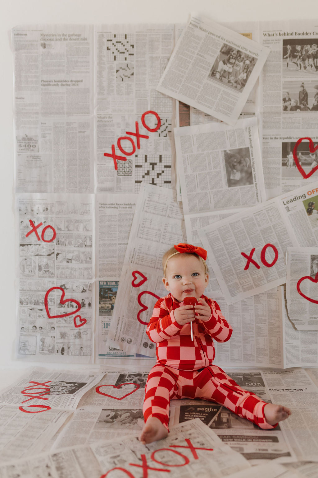 A baby in forever french baby's breathable Bamboo Zip Pajamas, XOXO edition, sits on the floor wearing a red and white checkered pattern. Newspaper pages with red "XO" and heart symbols create the backdrop. Holding a small item, the baby gazes directly into the camera.