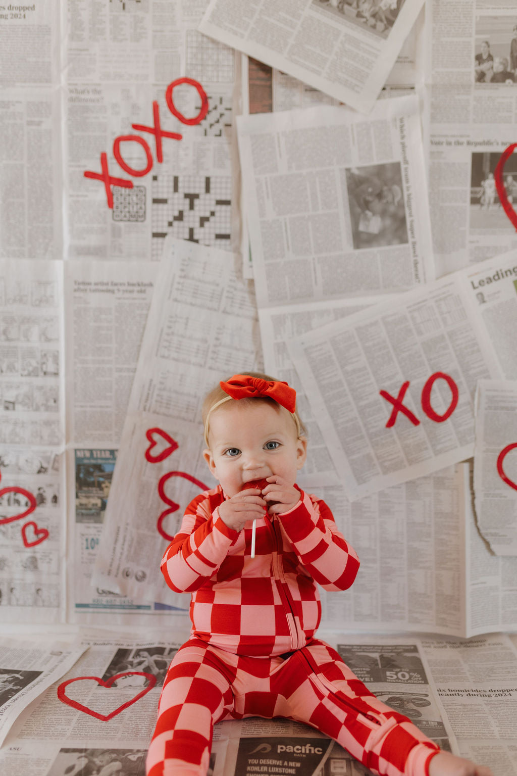 A small child sits on the floor in breathable Bamboo Zip Pajamas | XOXO by forever french baby. The outfit is red and white checkered with a red bow, and the background is covered in newspaper pages marked with red "XOXO" and hearts.