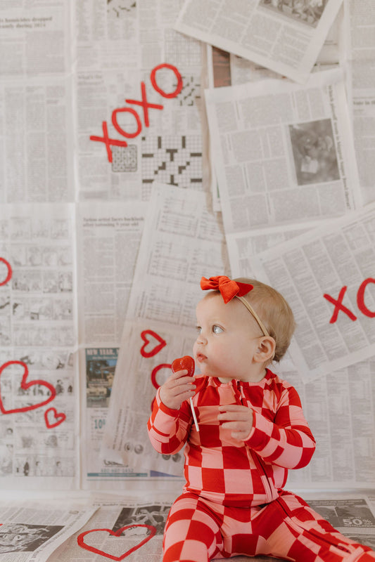 A baby in forever french baby's hypo-allergenic Bamboo Zip Pajamas | XOXO and a red headband holds a lollipop. A newspaper-covered wall displays red "XOXO" and hearts behind them.