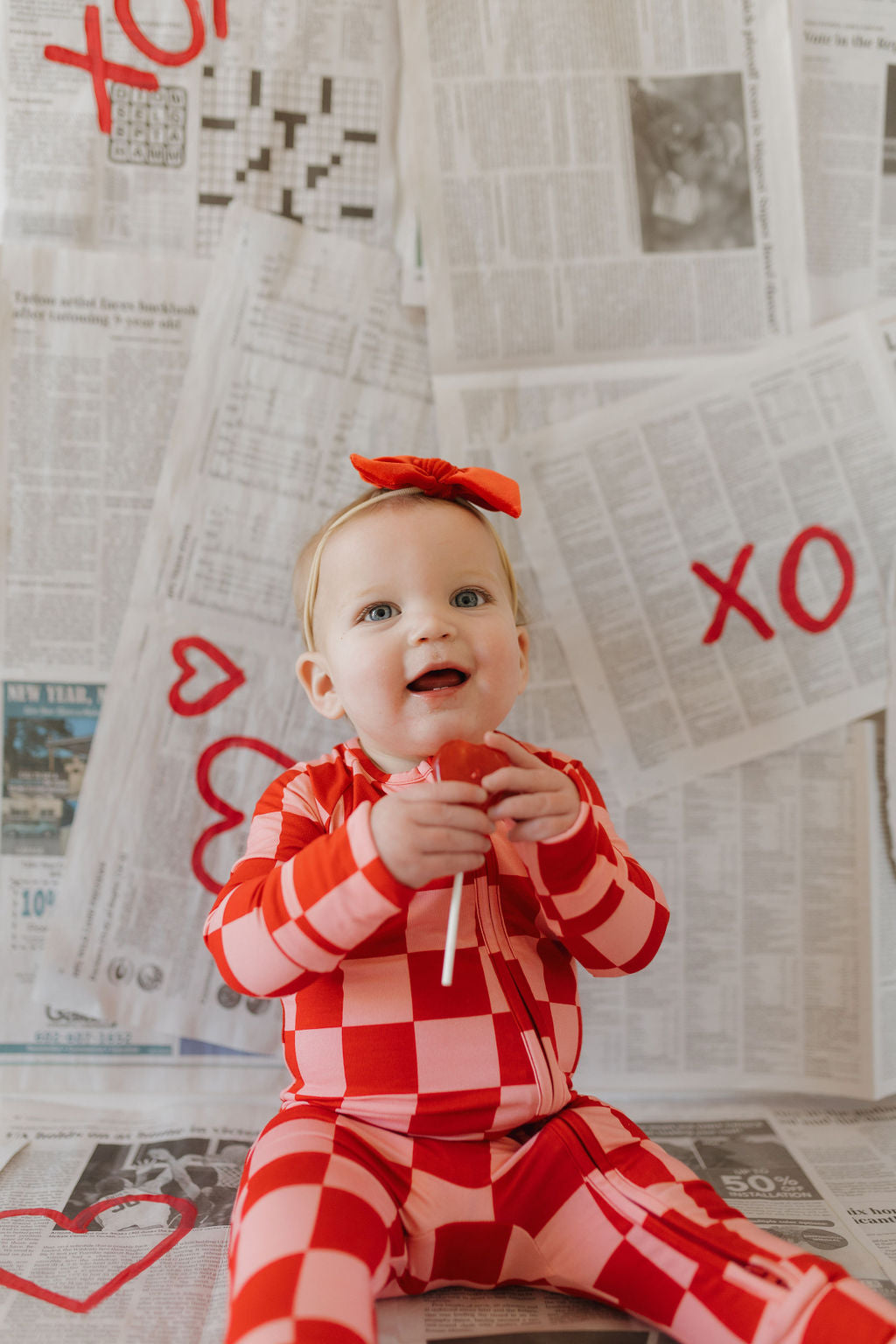 A baby in breathable, hypo-allergenic Bamboo Zip Pajamas by forever french baby and a matching headband sits on a newspaper-covered floor, holding a red heart lollipop. The background features "XO" and heart shapes painted in red on the newspapers.