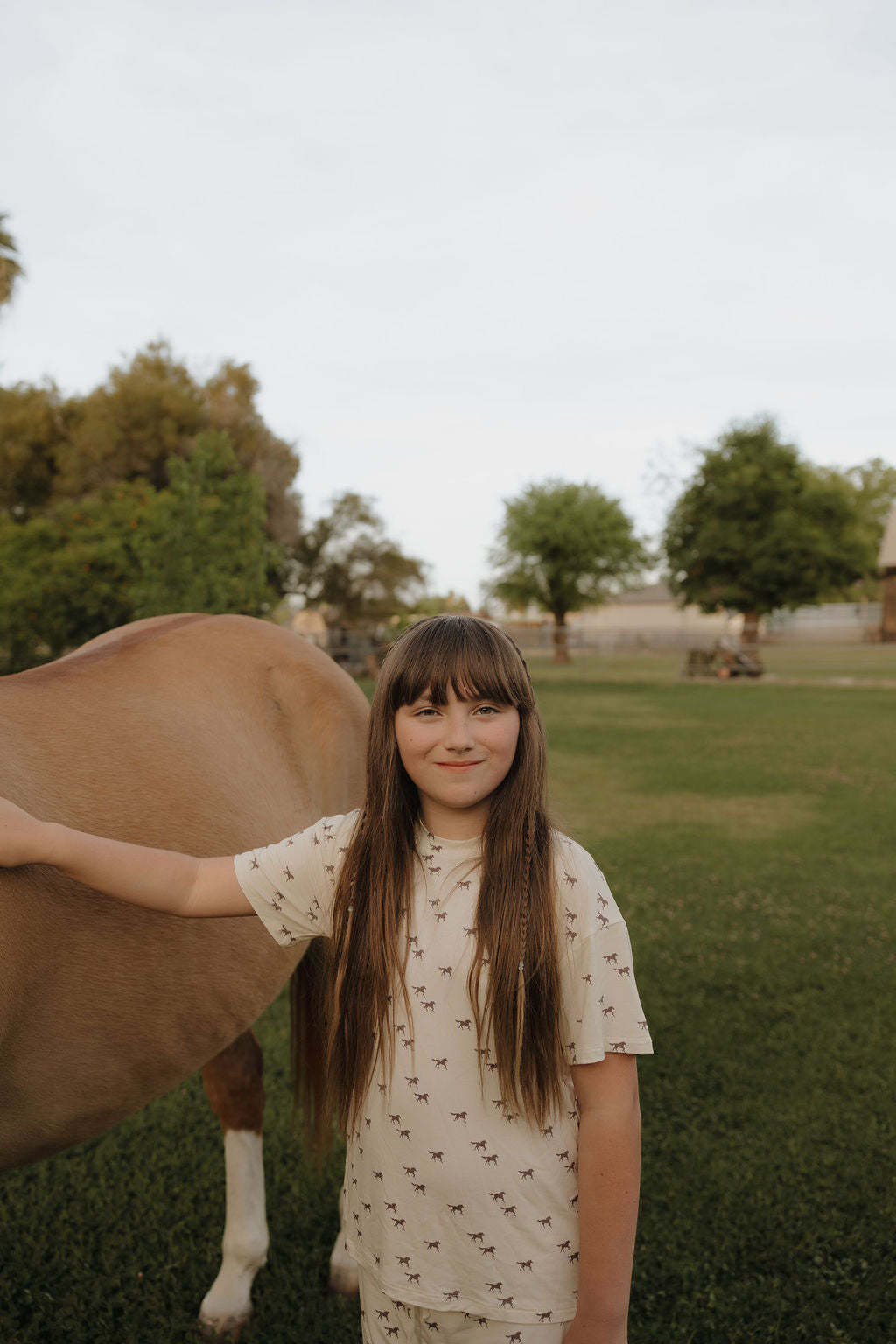 A smiling young girl with long brown hair wears the forever french baby Girl's Bamboo Short Set | Wild Horses, standing on green grass beside a horse. Trees and a fence appear in the background.