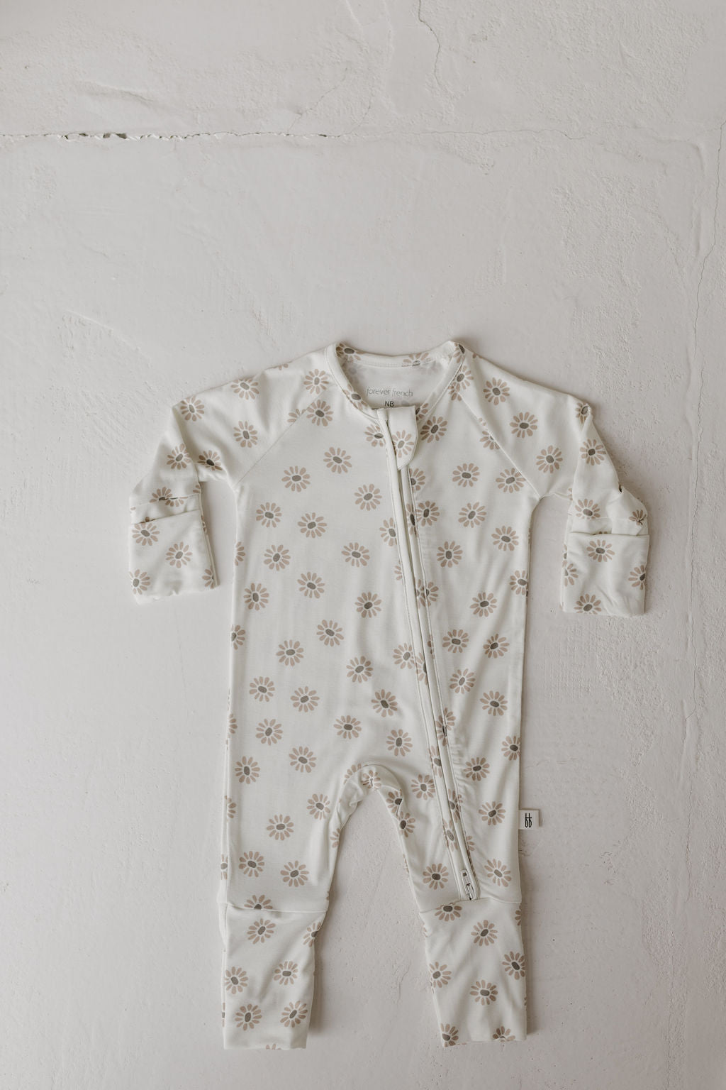 A "Bamboo Zip Pajamas | Daisy Fields" by forever french baby, features a white baby onesie with long sleeves and a front zipper made from hypo-allergenic fabric. Decorated with light brown florals, this sleepwear is displayed on a textured white surface.