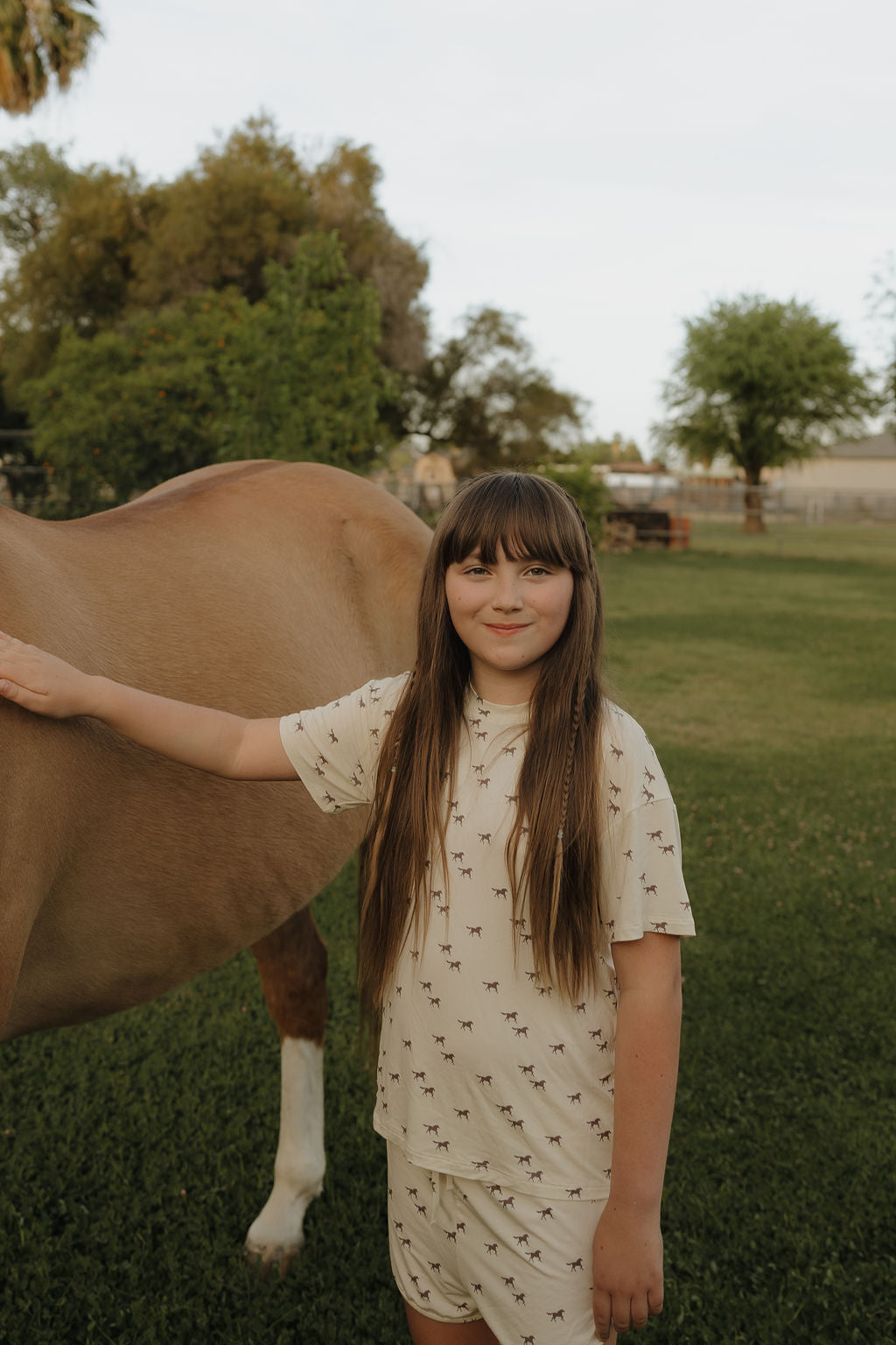 A young girl with long brown hair smiles at the camera, wearing the forever french baby Girl's Bamboo Short Set | Wild Horses. She stands on grass beside a brown horse, with trees and a fence in the background.