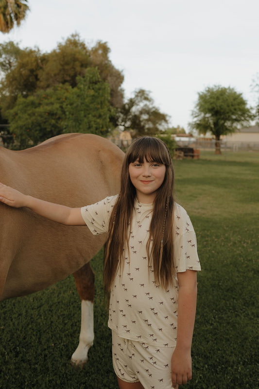 A young girl with long brown hair smiles at the camera, wearing the forever french baby Girl's Bamboo Short Set | Wild Horses. She stands on grass beside a brown horse, with trees and a fence in the background.