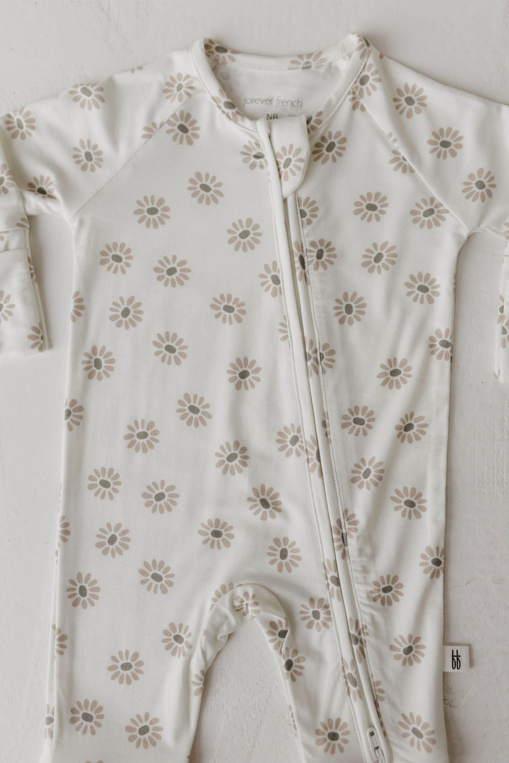 The Bamboo Zip Pajamas by forever french baby, in the Daisy Fields design, is a white, long-sleeve onesie with a light brown floral pattern. It's hypo-allergenic with a neck-to-leg zip closure and features a fabric label at the bottom right.