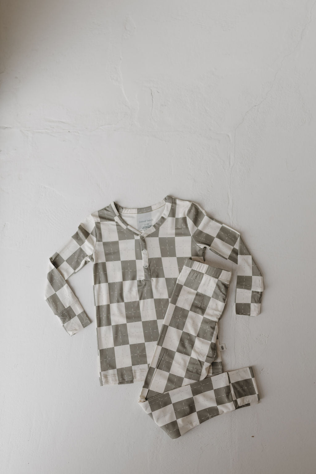 The "Bamboo Two Piece Pajamas | Oat & Sage" by forever french baby, featuring a breathable fabric with a checkered gray and white pattern, includes a long-sleeve top and matching pants displayed on a light, textured surface.