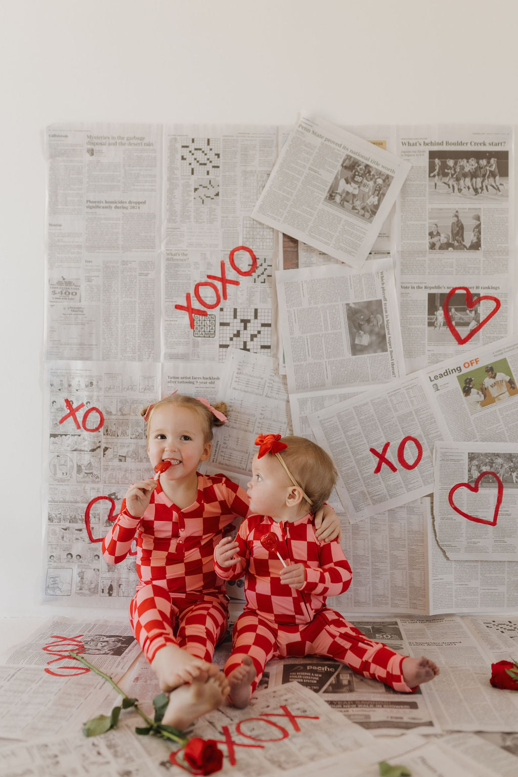 Two toddlers in forever french baby Bamboo Two Piece Pajamas | XOXO sit on a newspaper-covered floor, surrounded by red roses and paint writing "XOXO" alongside hearts. They hold lollipops, have red flowers in their hair, and enjoy the comfort of hypo-allergenic clothing.