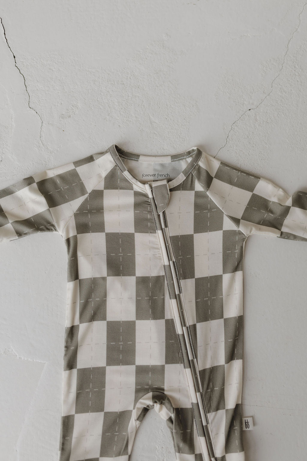 The Bamboo Zip Pajamas by forever french baby, adorned with a large beige and green checkered pattern, are crafted from hypo-allergenic bamboo fabric. This minimalist infant onesie features a central zipper and is displayed flat on a cracked white textured surface.