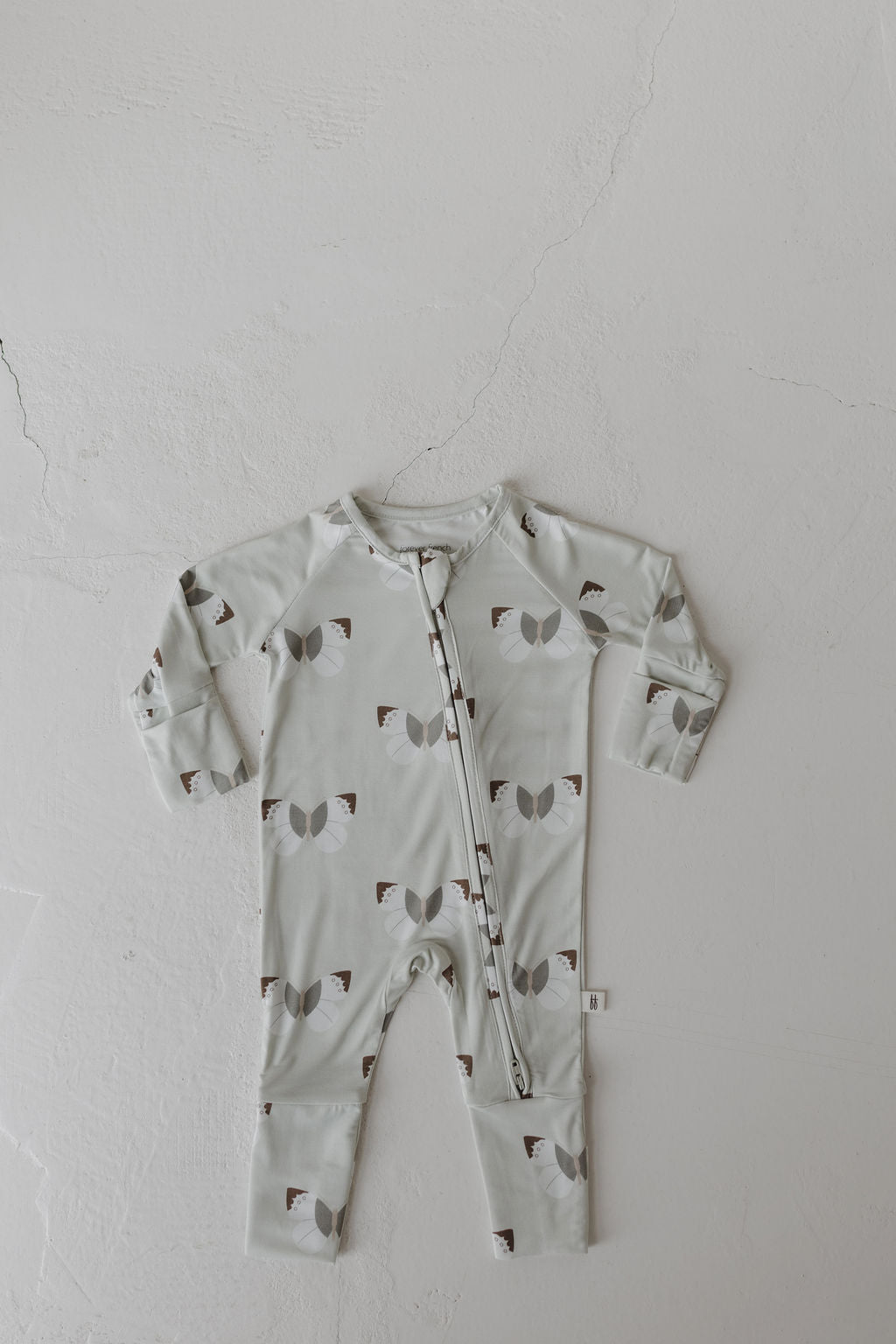 The Bamboo Zip Pajamas by forever french baby, featuring a front zipper, is laid flat on a cracked white surface. This hypo-allergenic onesie is light gray, adorned with abstract shapes in darker gray and brown, resembling bows or hourglasses.